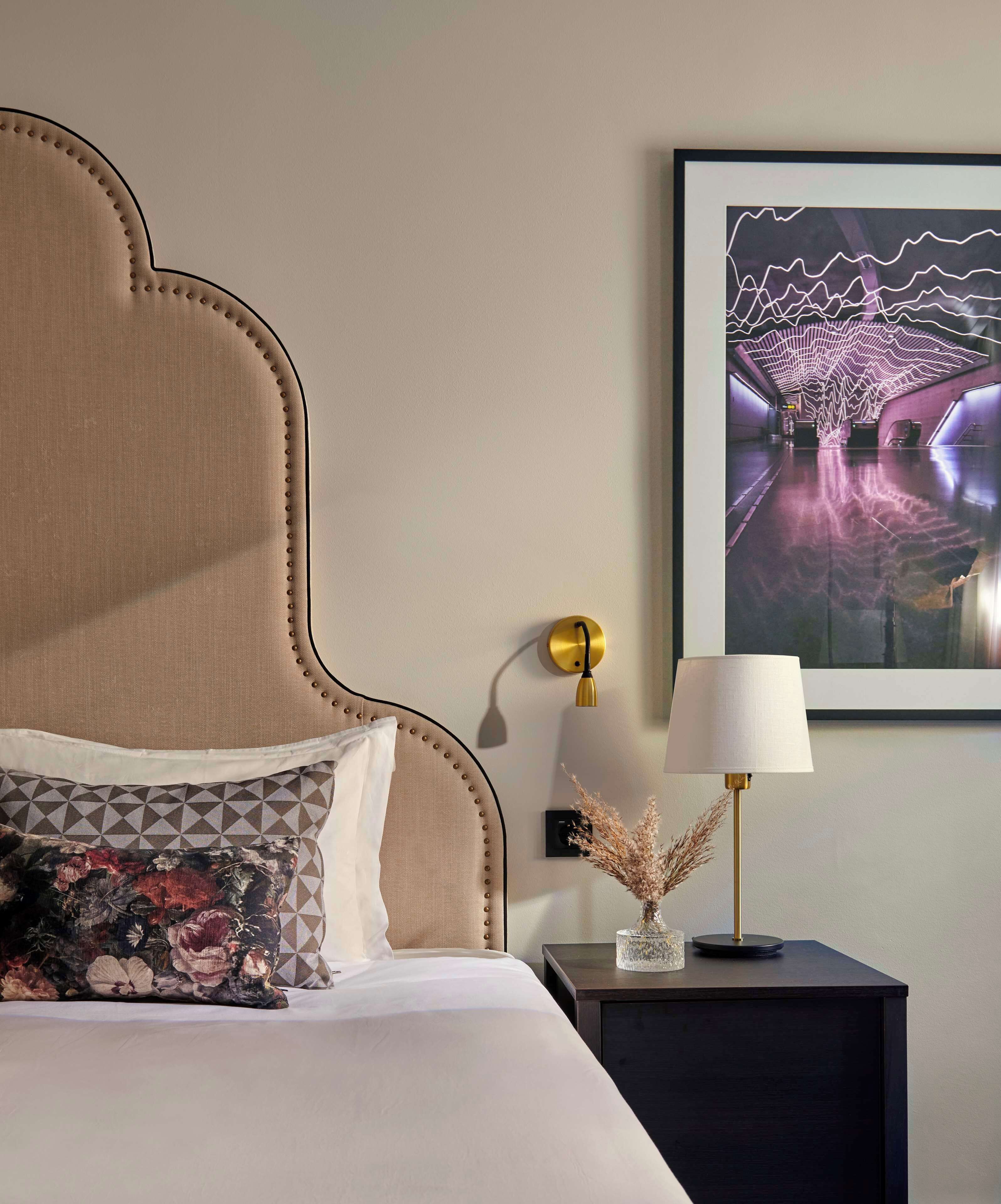 A bedroom with a large bed, a nightstand, a lamp, a framed picture, and a wall-mounted light.