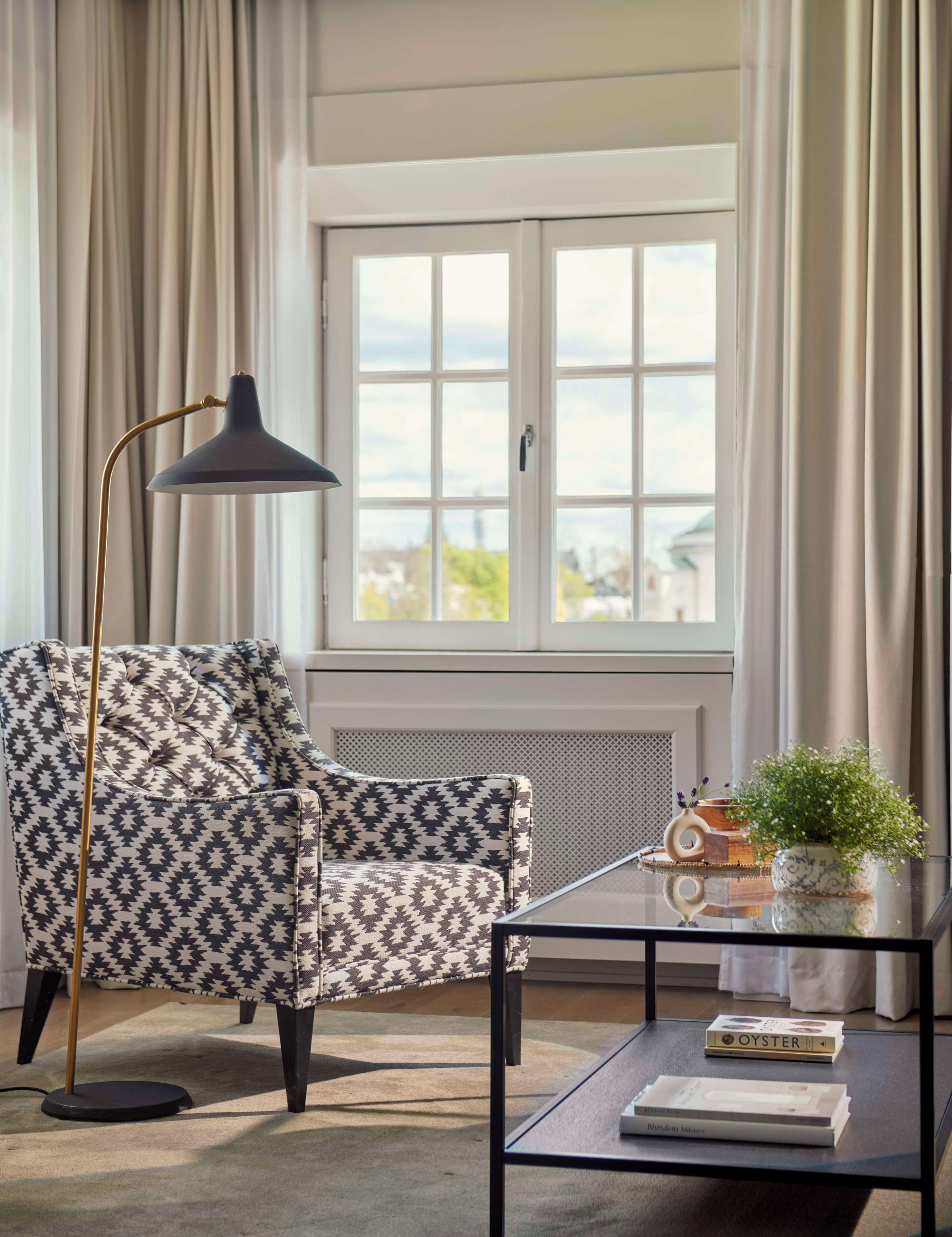 A modern living room with a patterned armchair, a floor lamp, a glass coffee table with books and a potted plant, and a view through a large window with curtains.