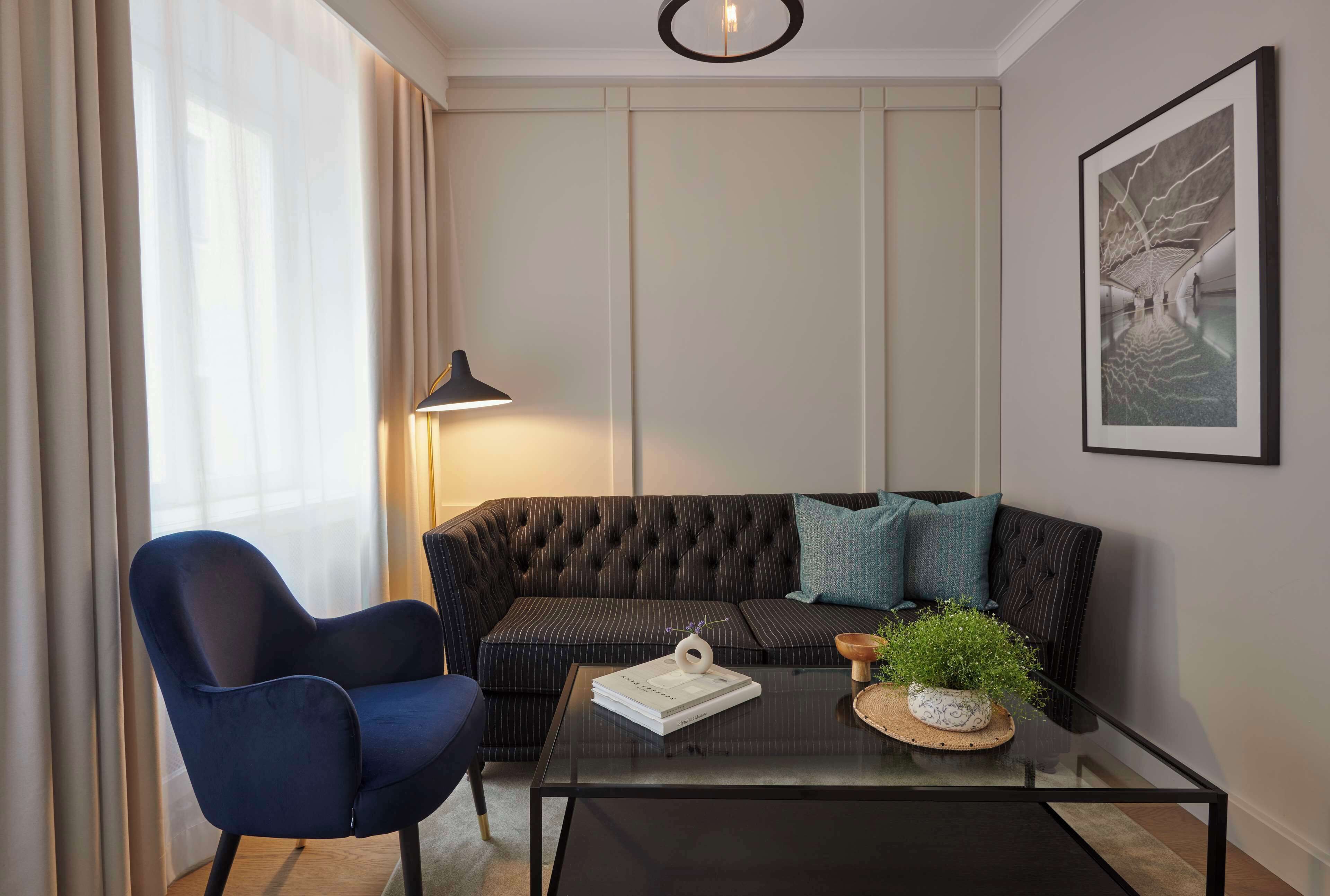 A modern living room with a dark upholstered sofa, a blue armchair, a glass coffee table, and a floor lamp.