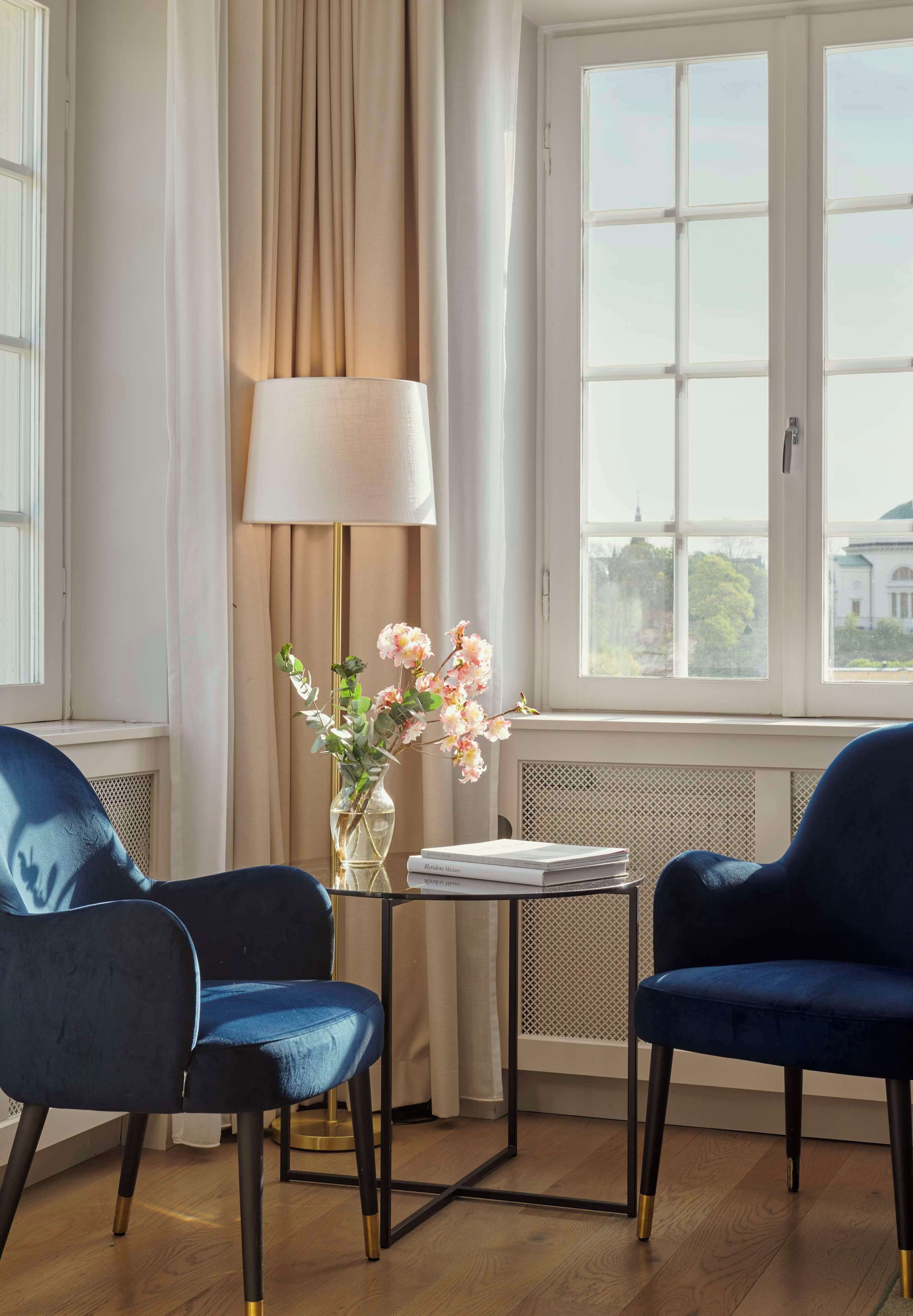 Quarto com duas cadeiras azuis, uma mesa com abajur e um vaso de flores, e uma janela com vista para um edifício.