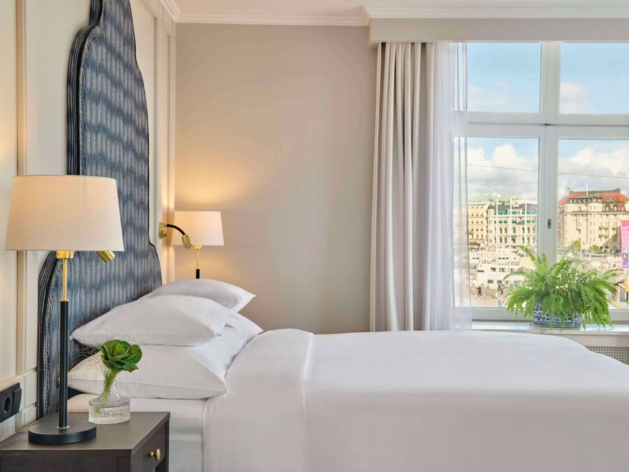 Ein Hotelzimmer mit einem großen Bett, einem Nachttisch mit Lampe, einer Topfpflanze und Blick auf die Stadt durch ein großes Fenster.