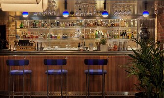 Zona de bar con varias estanterías llenas de botellas, vasos y utensilios. Hay tres taburetes azules y una planta en maceta.