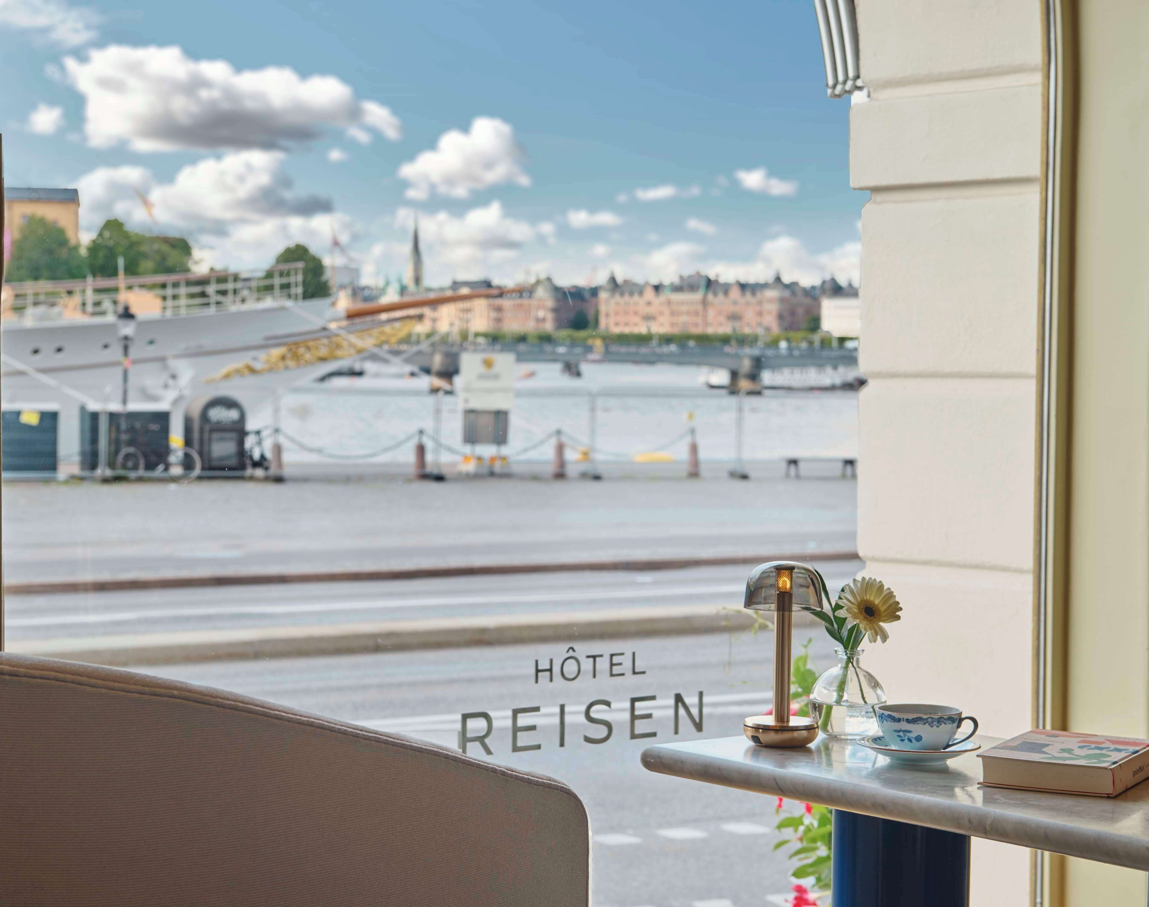 A view from a hotel room with a table displaying a vase with flowers, a cup, and a book. The room has a window with a view of a waterfront and a bridge. The text "HOTEL REISEN" is visible on the window.
