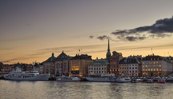 Un paisaje urbano frente al mar con edificios históricos, incluyendo una imponente aguja de iglesia, y varios barcos amarrados en el puerto.