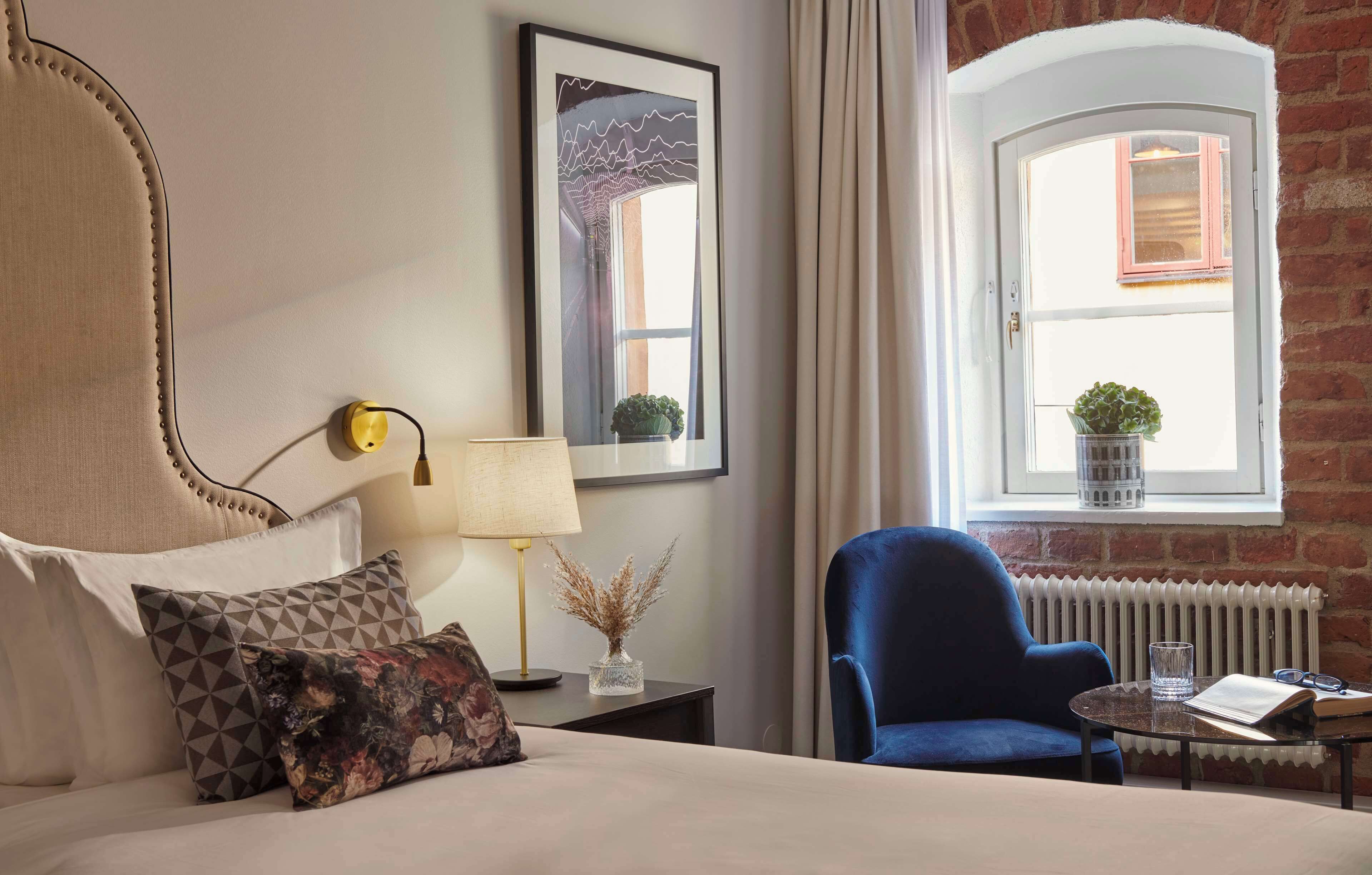 A hotel room with a bed, a blue armchair, a small table with a book and glasses, a lamp, a framed picture, and a window with a view of a brick wall and a window with a plant.