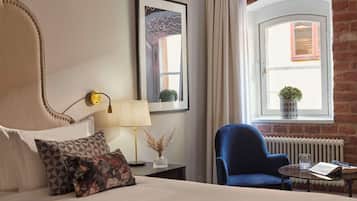 A hotel room with a bed, a blue armchair, a small table with a book and glasses, a lamp, a framed picture, and a window with a view of a brick wall and a window with a plant.