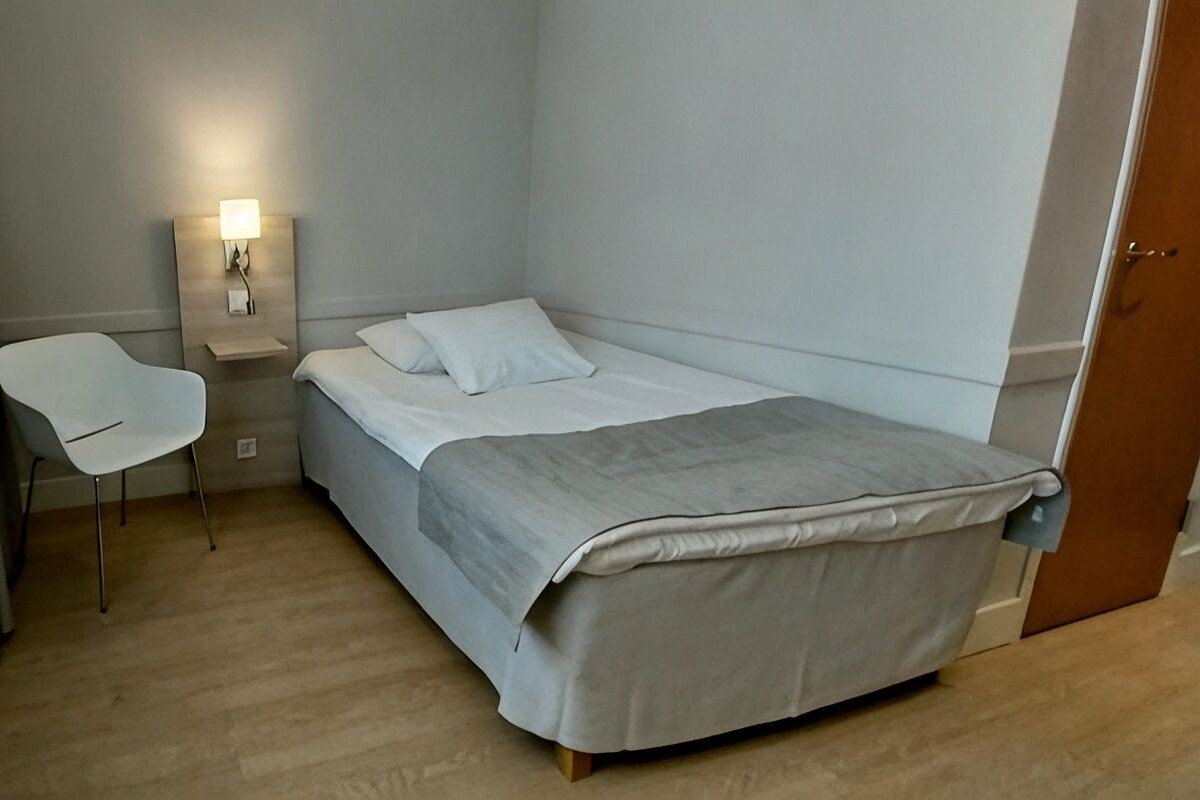 A single bed with a grey blanket, a bedside table with a lamp, and a white chair in a room with wooden flooring.