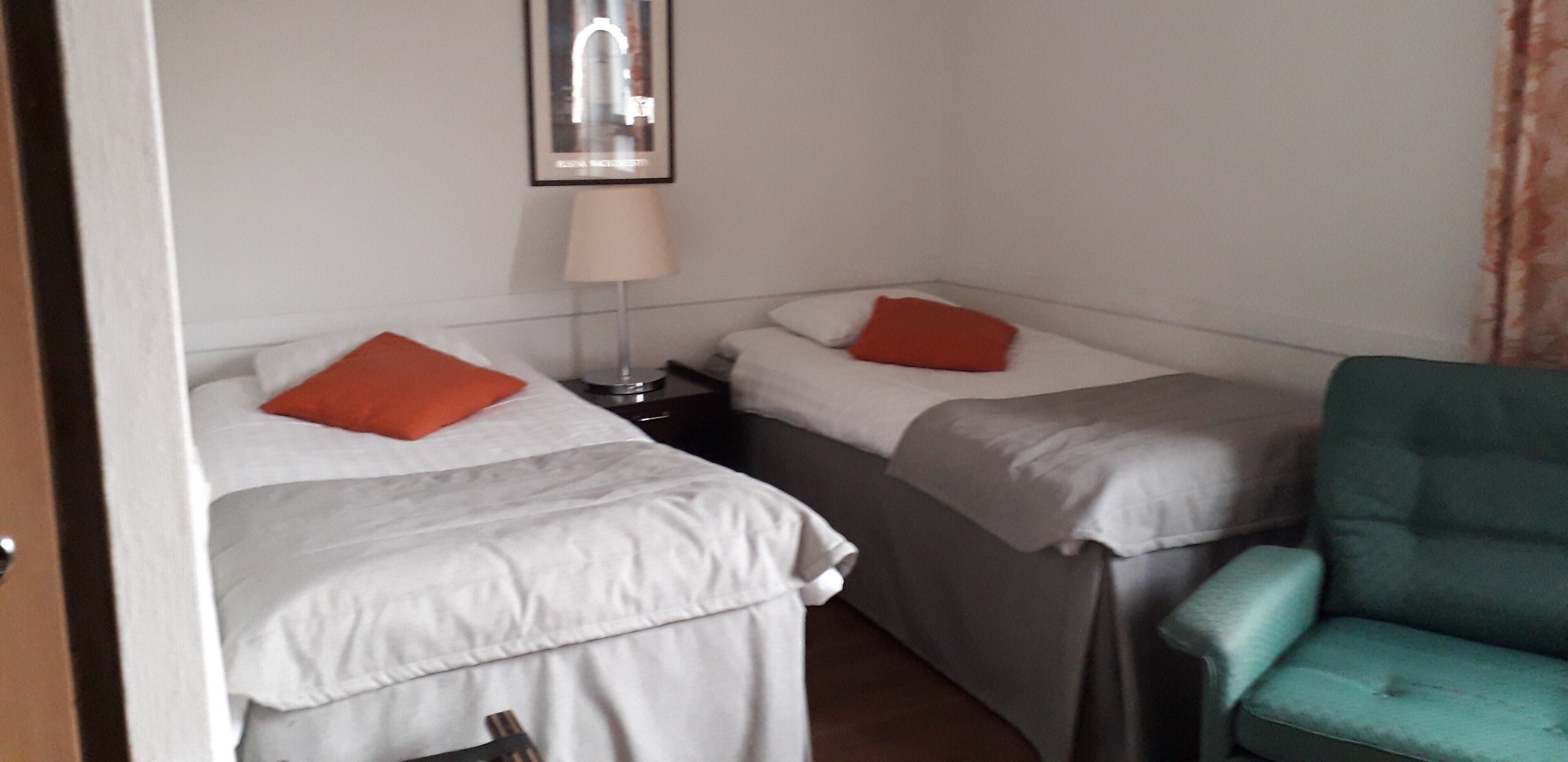 Ein Zimmer mit zwei Betten, jeweils mit einem Kissen und einer Lampe dazwischen. Eine grüne Sitzgelegenheit ist rechts zu sehen.