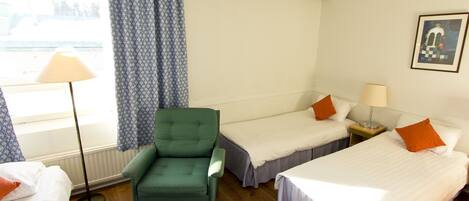 A hotel room with two single beds, a green armchair, a small table, and a window with curtains.