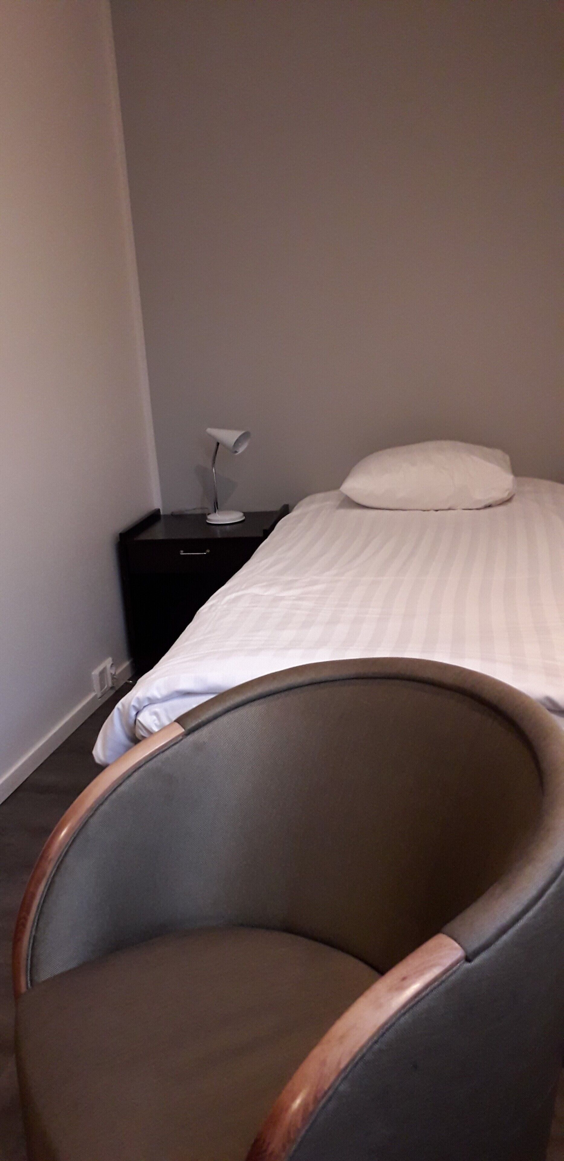 Quarto de hotel com uma cama de solteiro, mesa de cabeceira com abajur e uma cadeira.