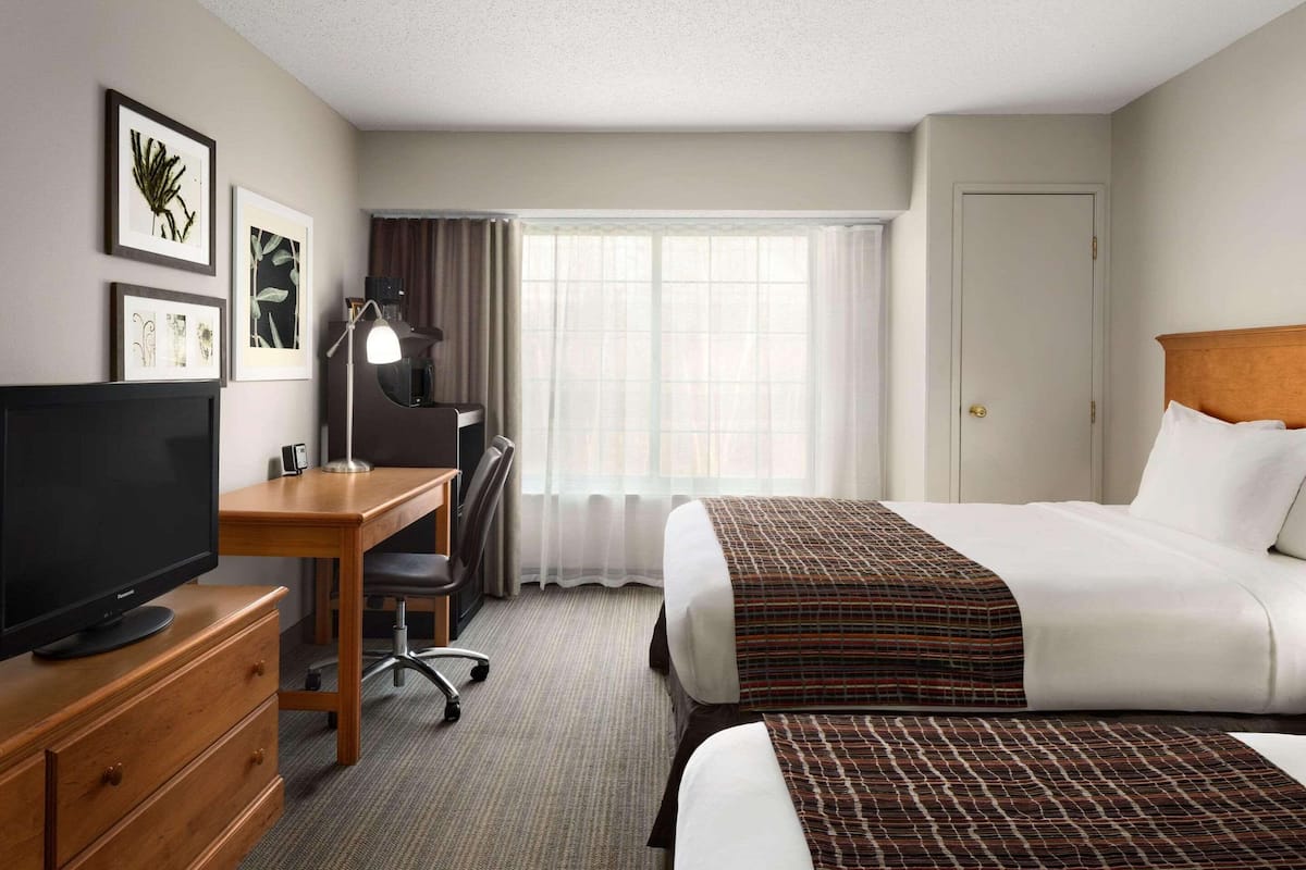 A hotel room with a bed, a desk with a computer, a TV, and a window with curtains.