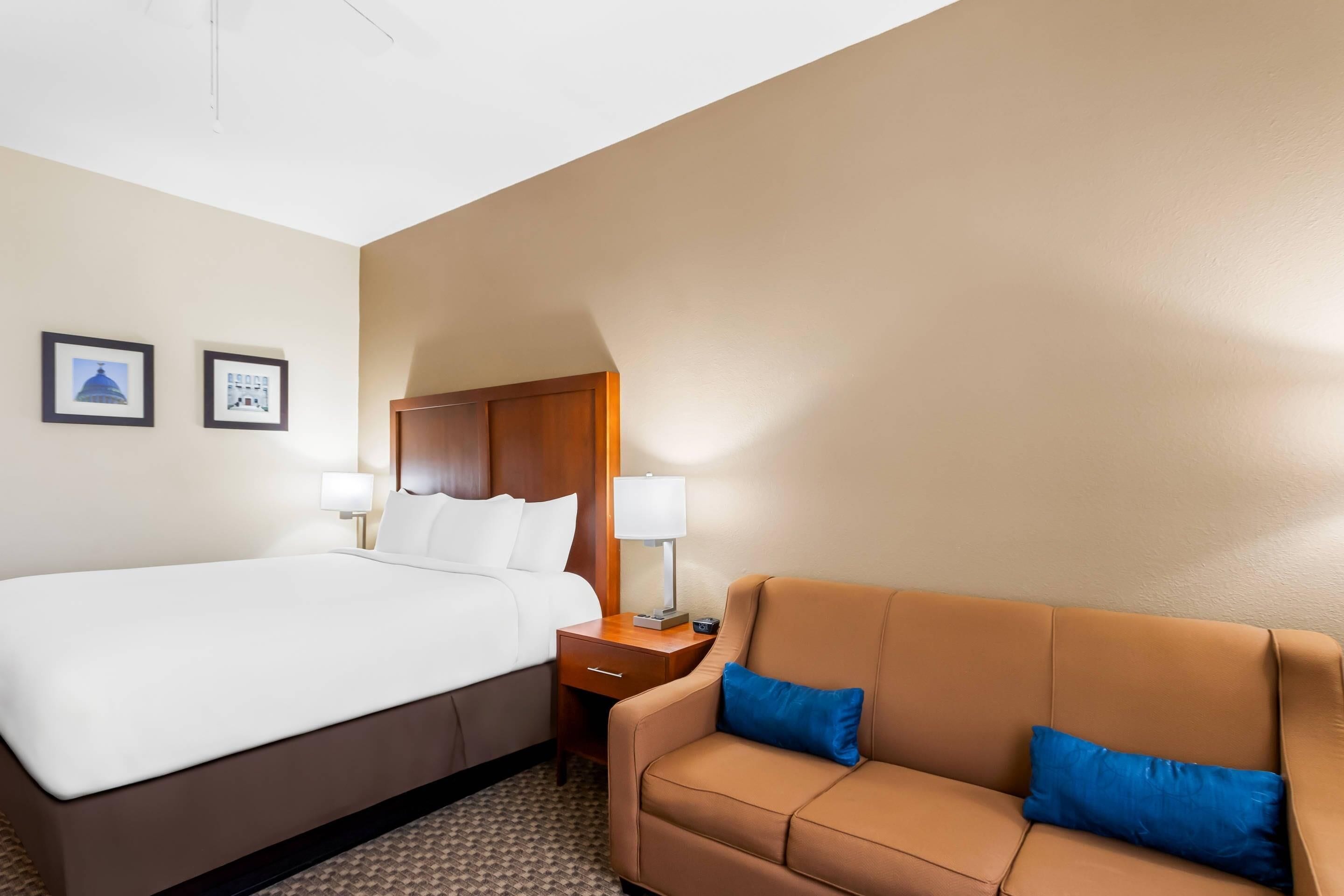 A hotel room with a bed, a brown sofa, two wall-mounted lamps, and framed pictures on the wall.