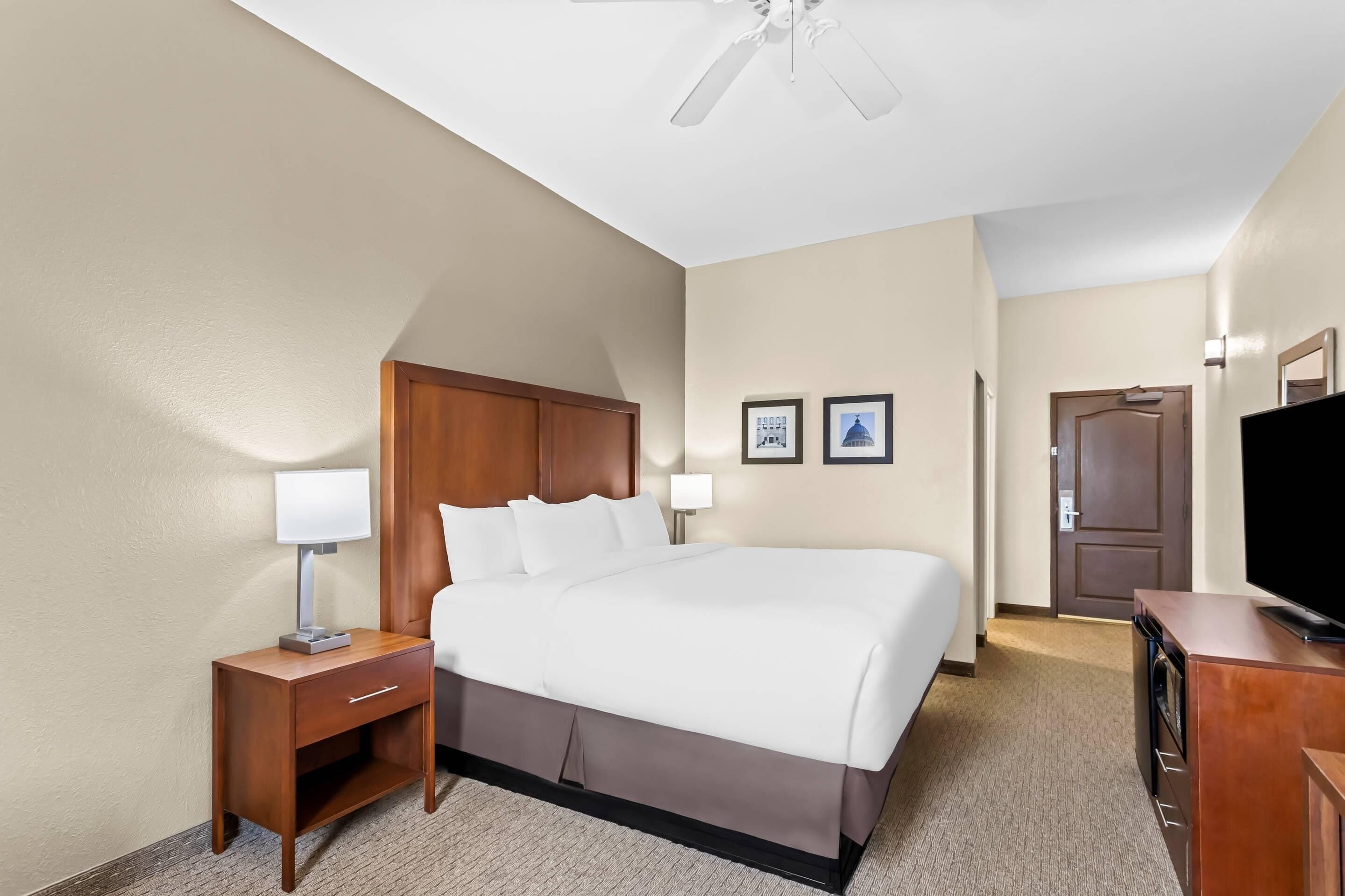 Une chambre d’hôtel avec un lit, une table de chevet avec une lampe, une télévision et un ventilateur de plafond.