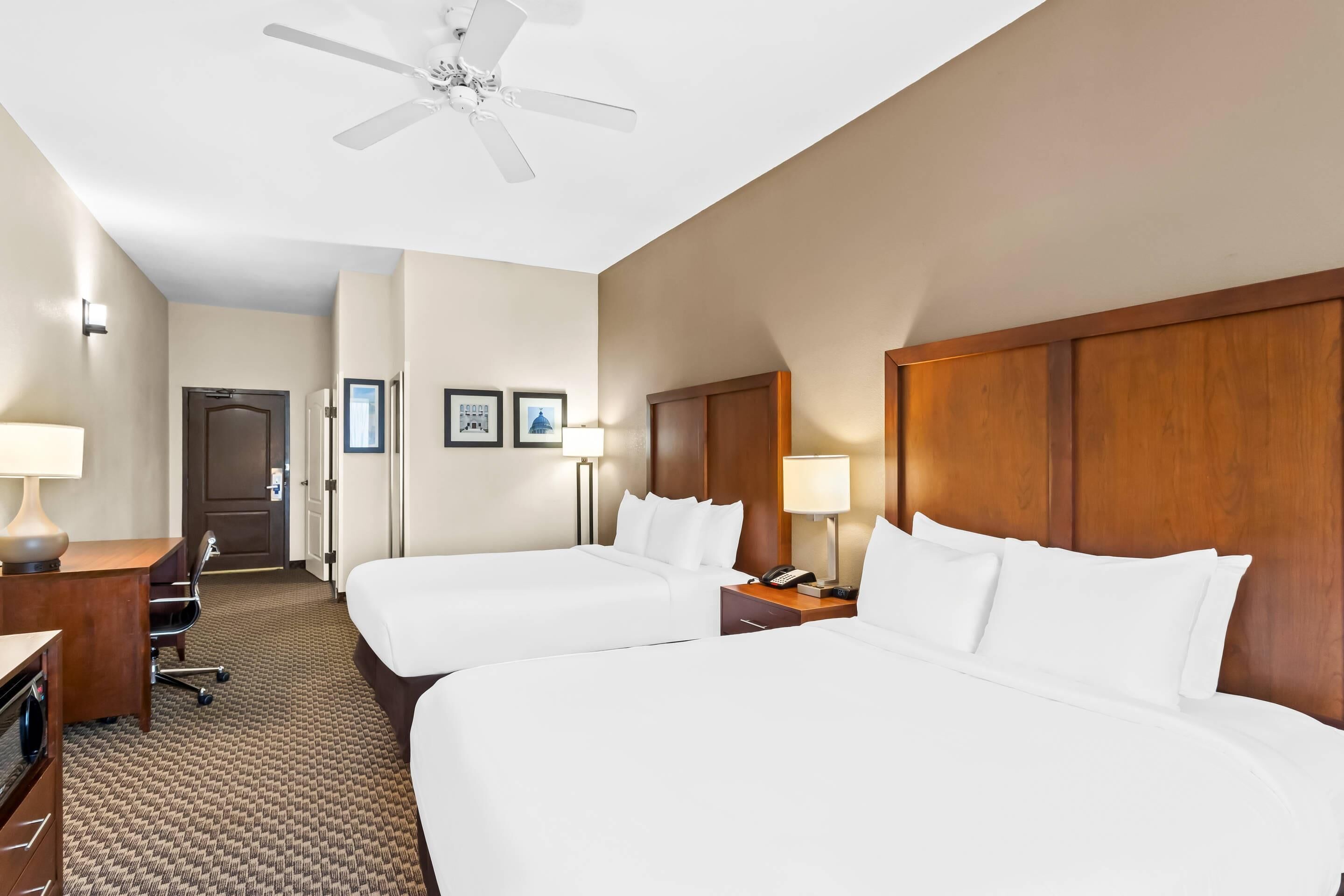 A hotel room with two beds, a desk, a chair, a desk lamp, a nightstand, a phone, a ceiling fan, and framed pictures on the wall.