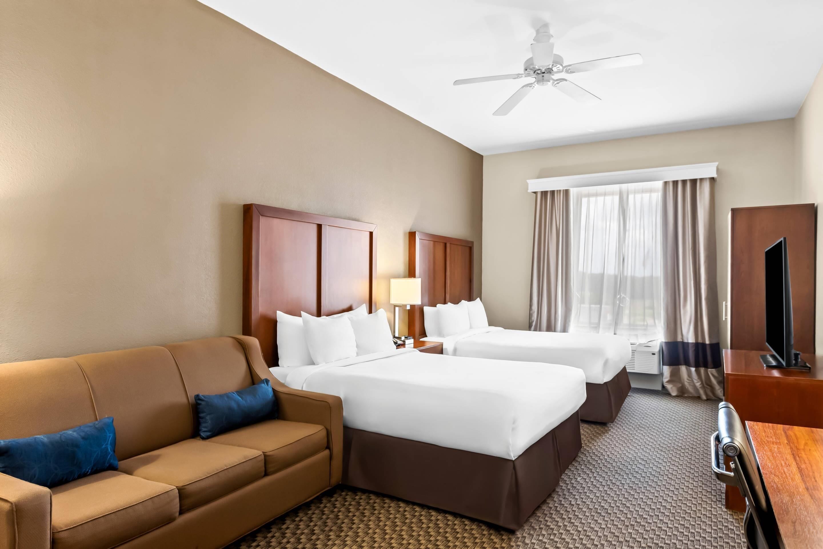 A hotel room with two beds, a sofa, a desk, a TV, and a ceiling fan.