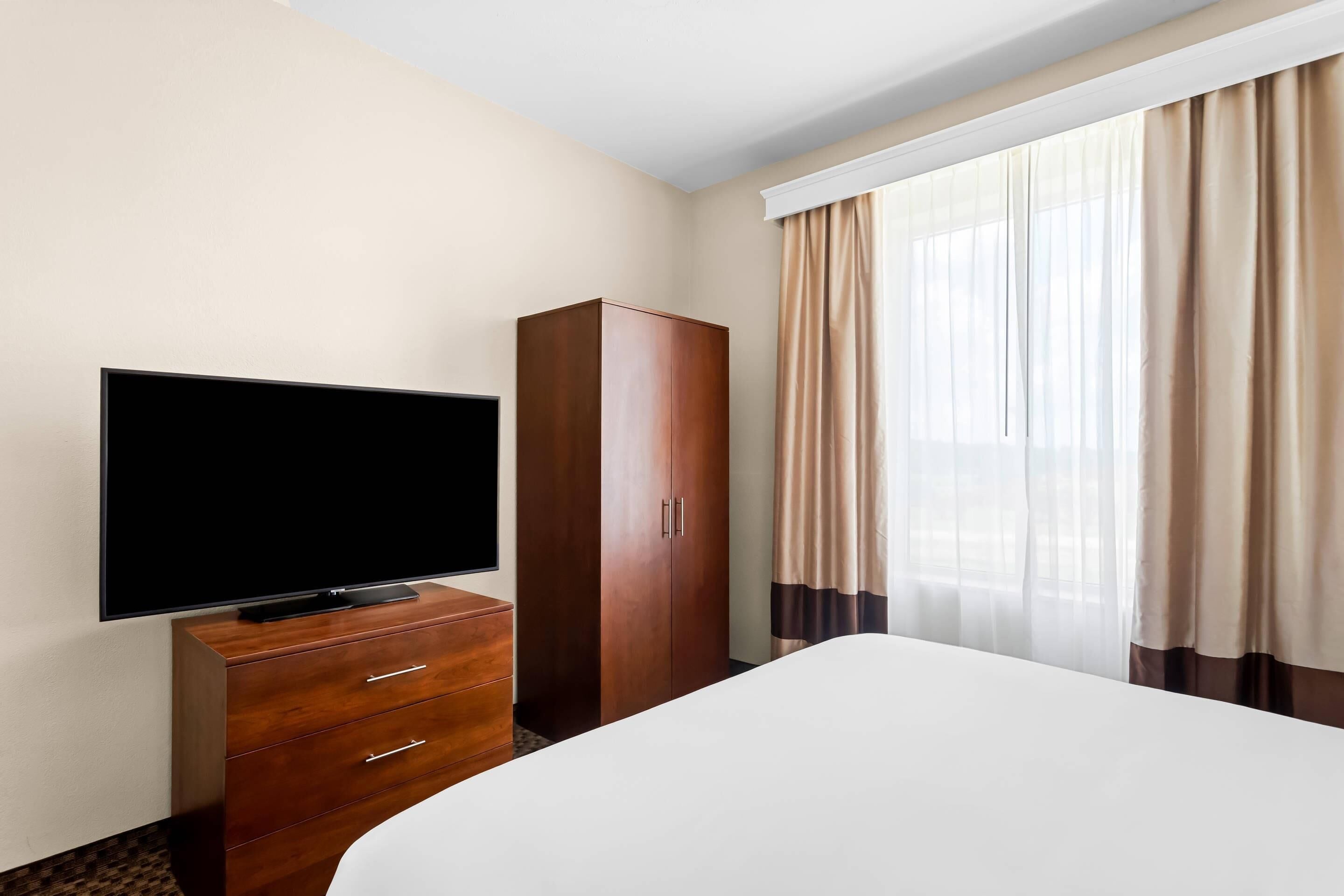 A hotel room with a bed, a TV on a wooden dresser, a wardrobe, and a window with curtains.