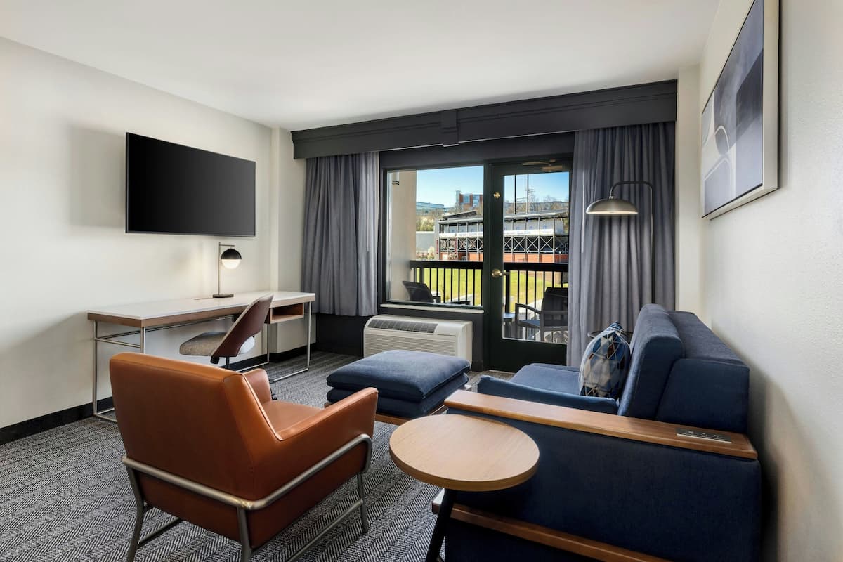 A hotel room with a flat-screen TV, a desk, a brown leather chair, a blue sofa, a small round table, and a balcony with a view of a building and a body of water.