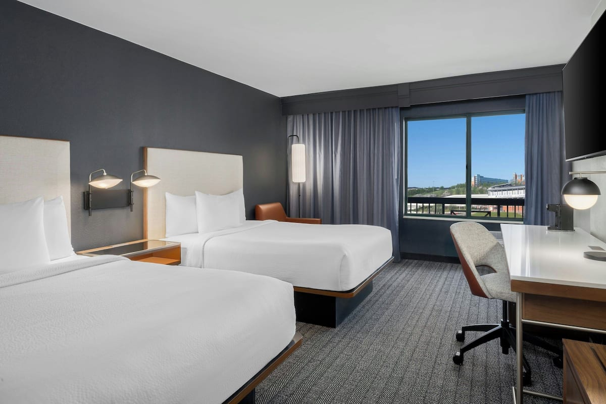 A hotel room with two beds, a desk, and a chair. There is a large window with curtains, and a view of the cityscape.