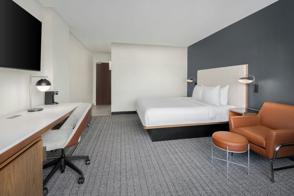A modern hotel room with a bed, a desk with a chair, a sofa, and a TV mounted on the wall.