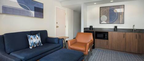 Quarto de hotel moderno com sofá, apoio para os pés, pequena mesa, poltrona de couro, cozinha compacta com micro-ondas e uma grande pintura abstrata na parede.