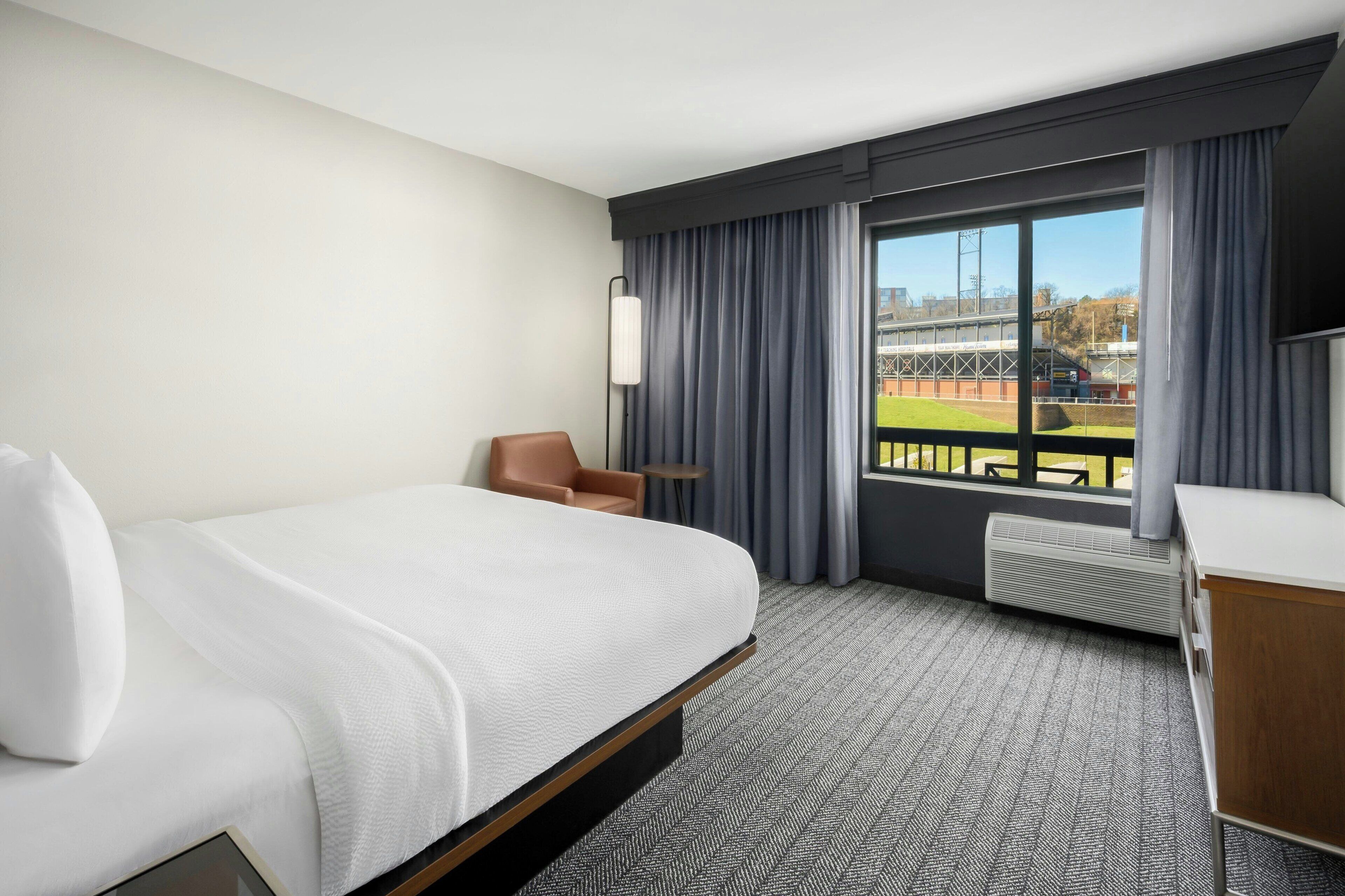 A hotel room with a large bed, a desk, a chair, and a view of a sports field through a large window.