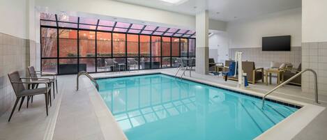 An indoor swimming pool with clear glass walls, a tiled floor, and a seating area with chairs and tables.