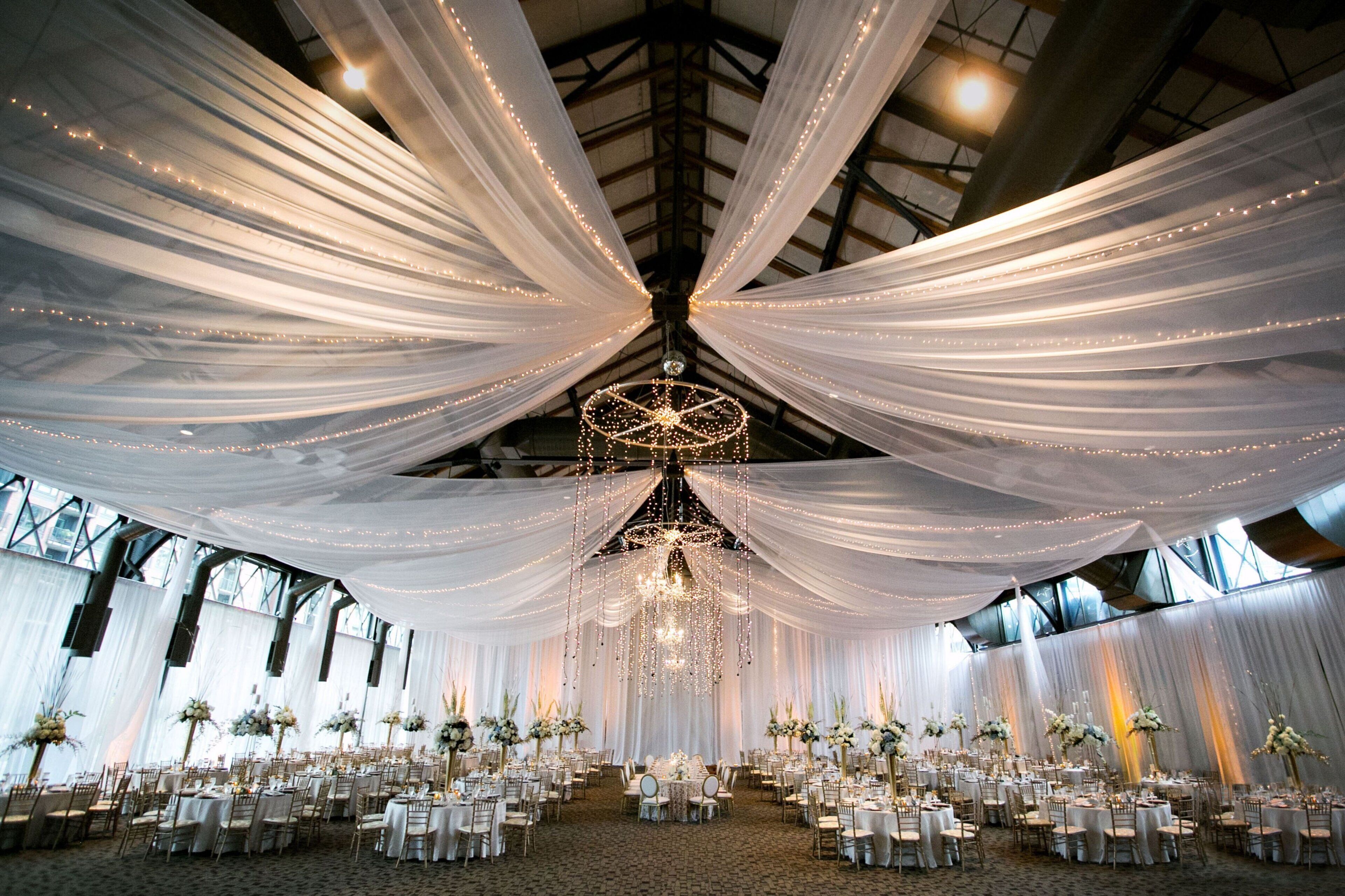 A large hall with draped white curtains, chandeliers, and tables set for a formal event.