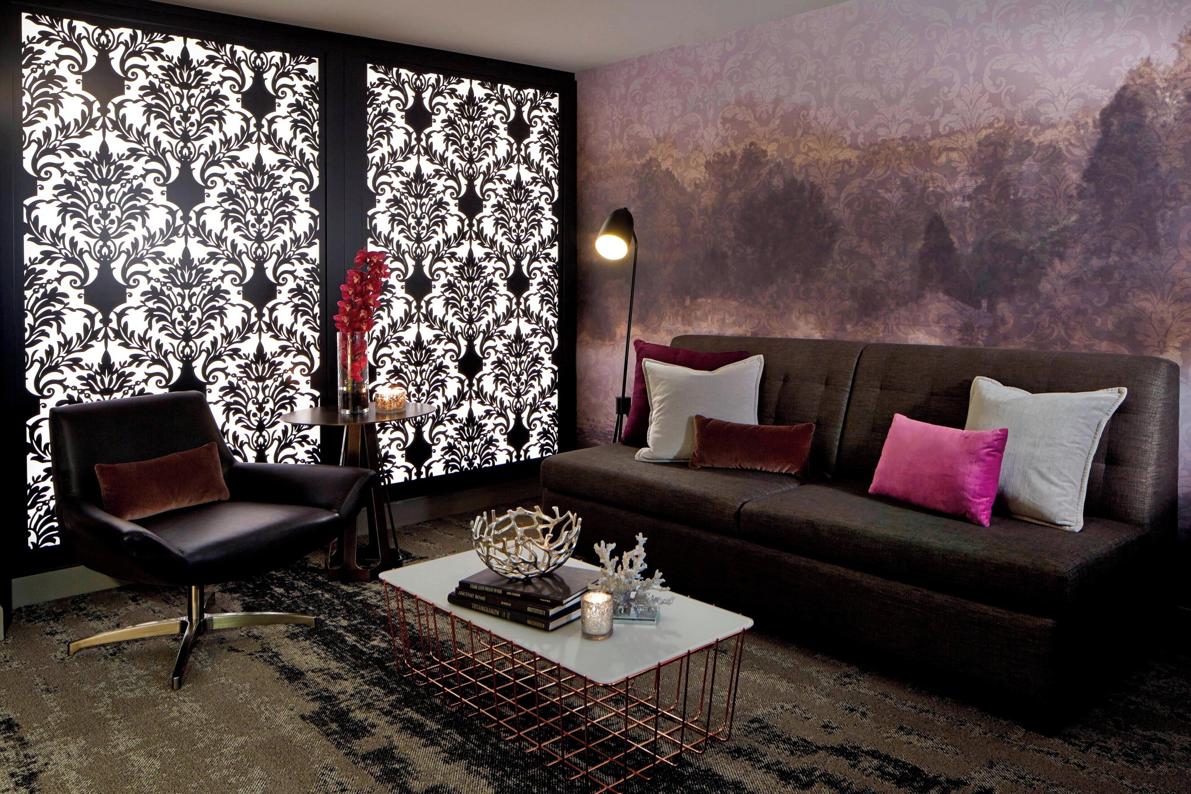 Een moderne woonkamer met een zwarte leren fauteuil, een bruine bank met meerdere kussens, een witte salontafel met decoratieve objecten, en een behang met een patroon.