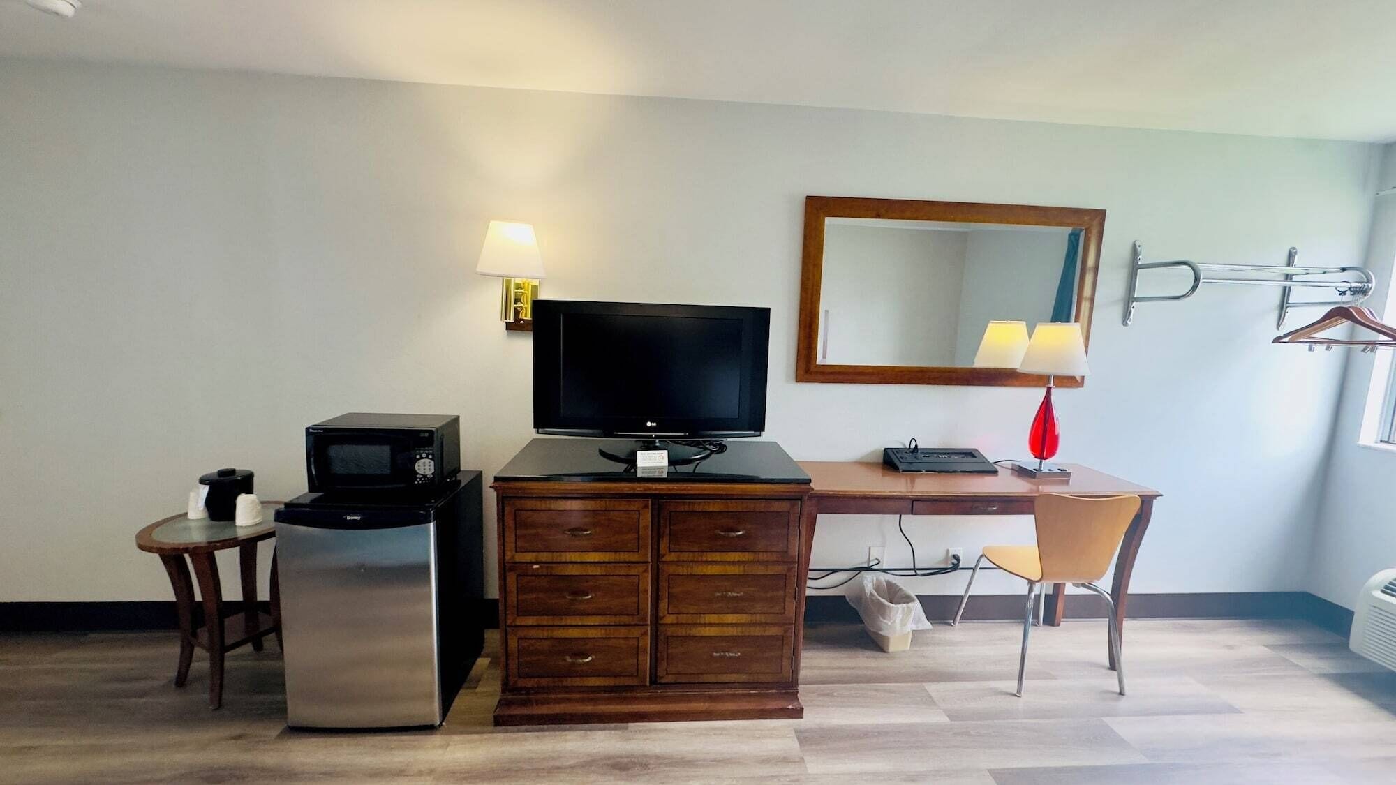 A hotel room with a wooden desk, a television, a microwave, a small table with a cup, and a chair.