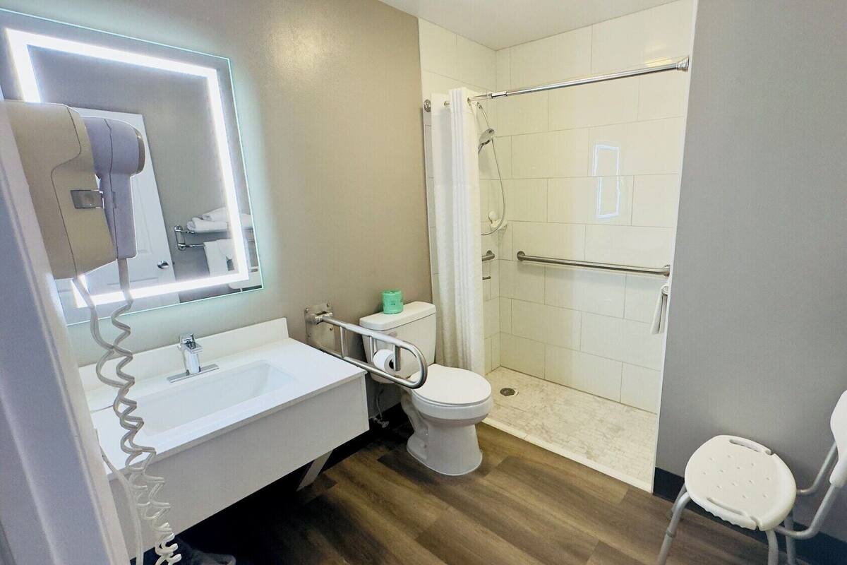 A modern bathroom with a white sink, toilet, and shower. There is a handrail in the shower, a towel rack, and a mirror above the sink.