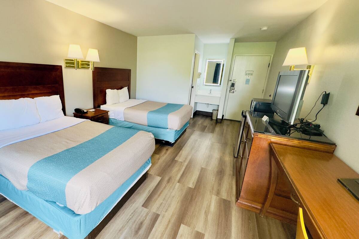 A hotel room with two beds, a wooden headboard, bedside lamps, a desk with a TV, and a bathroom visible through an open door.