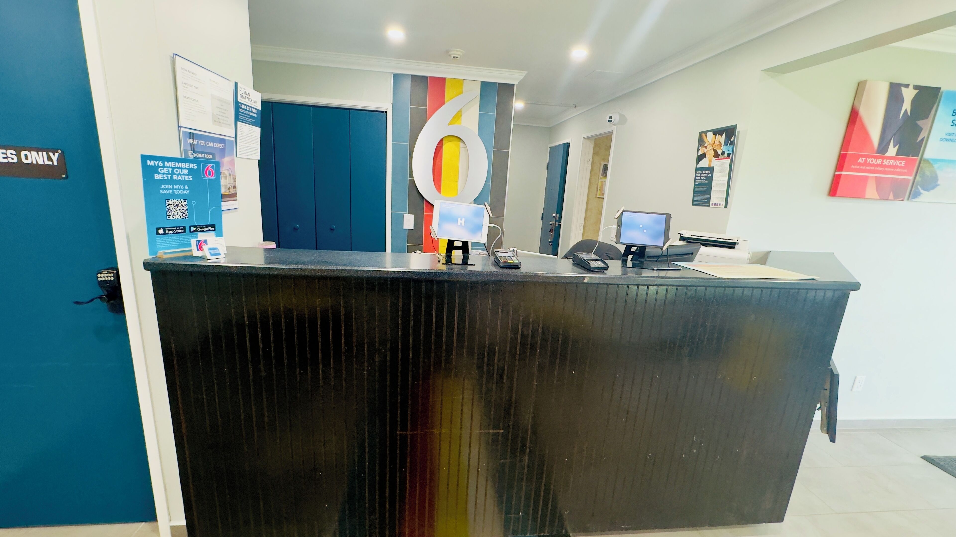 Un mostrador de recepción con un gran letrero con el número '6', una computadora y un teléfono. Hay carteles en la pared y una puerta azul con un letrero que dice 'RESERVES ONLY'.