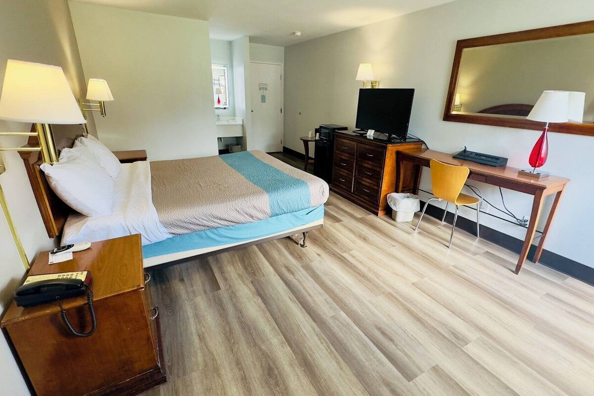 A hotel room with a bed, bedside table, lamp, desk, chair, TV, and a mirror.