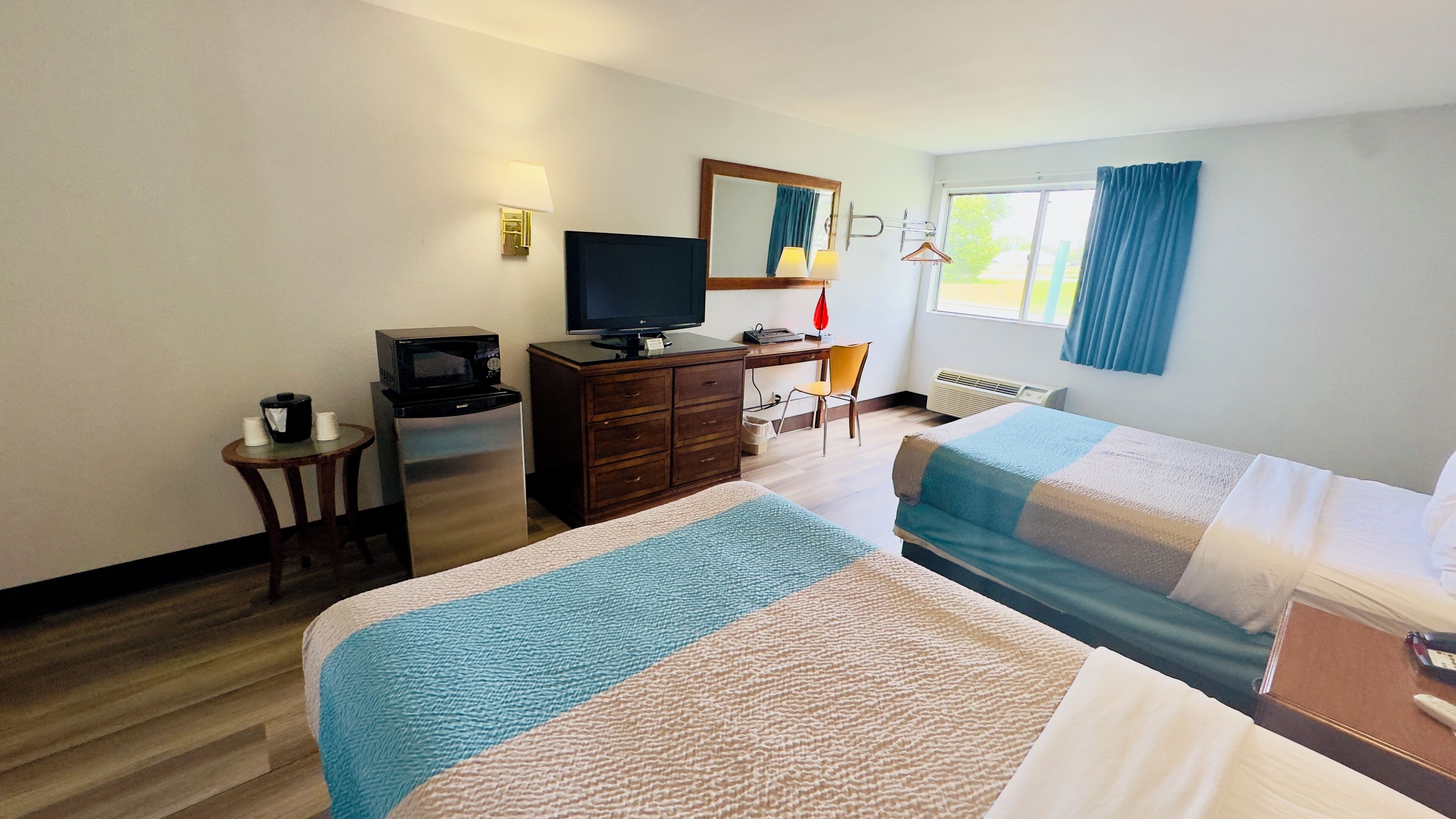 Habitación de hotel con dos camas, un escritorio con televisor, una mesita con cafetera, un espejo y una ventana con cortinas.