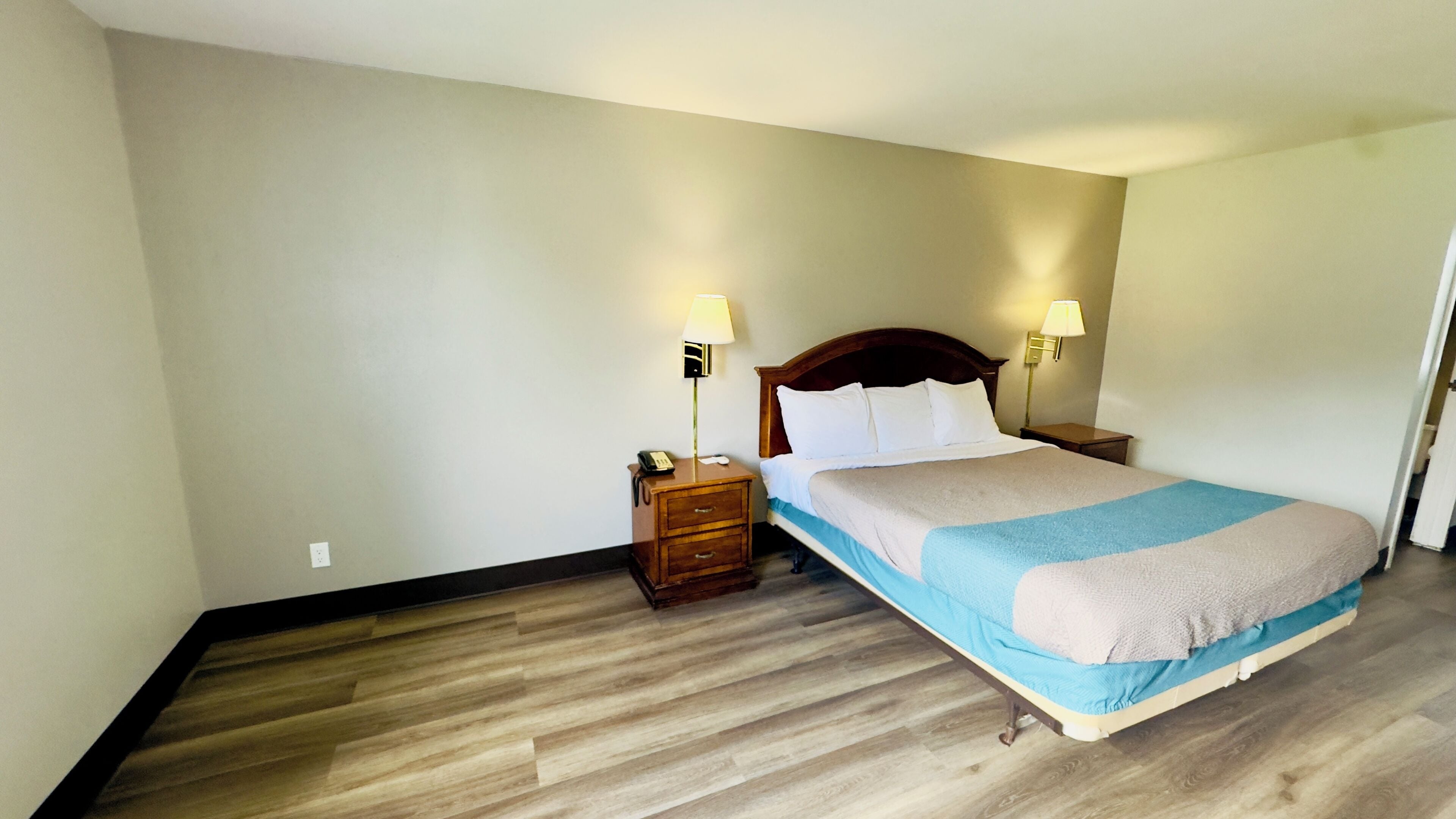 A hotel room with a wooden floor, a bed with a blue and beige bedspread, two bedside lamps, and a nightstand with a phone.