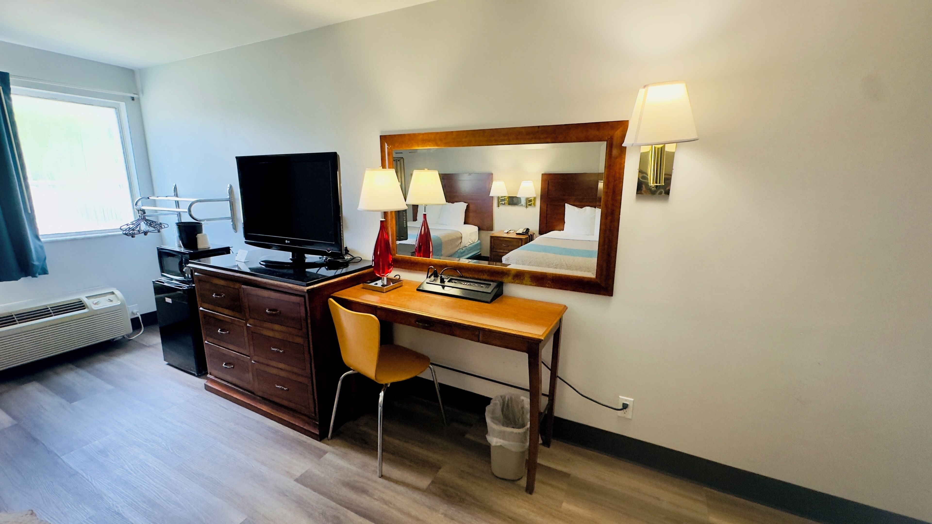 Une chambre d’hôtel avec un bureau en bois, une chaise, une télévision, un miroir et une fenêtre avec des rideaux.