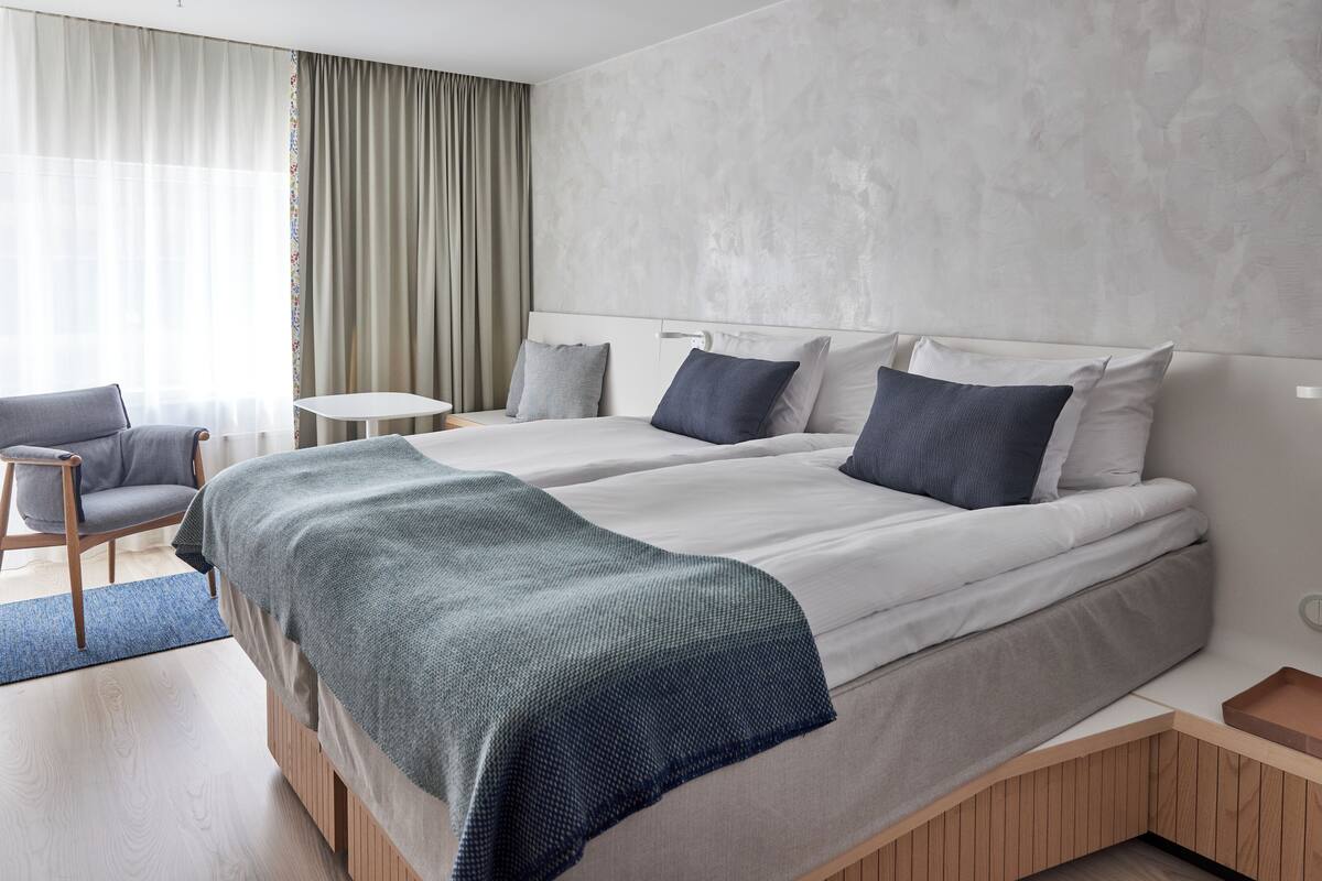 A modern hotel room with two beds, a small table, a chair, and a view of the outdoors.