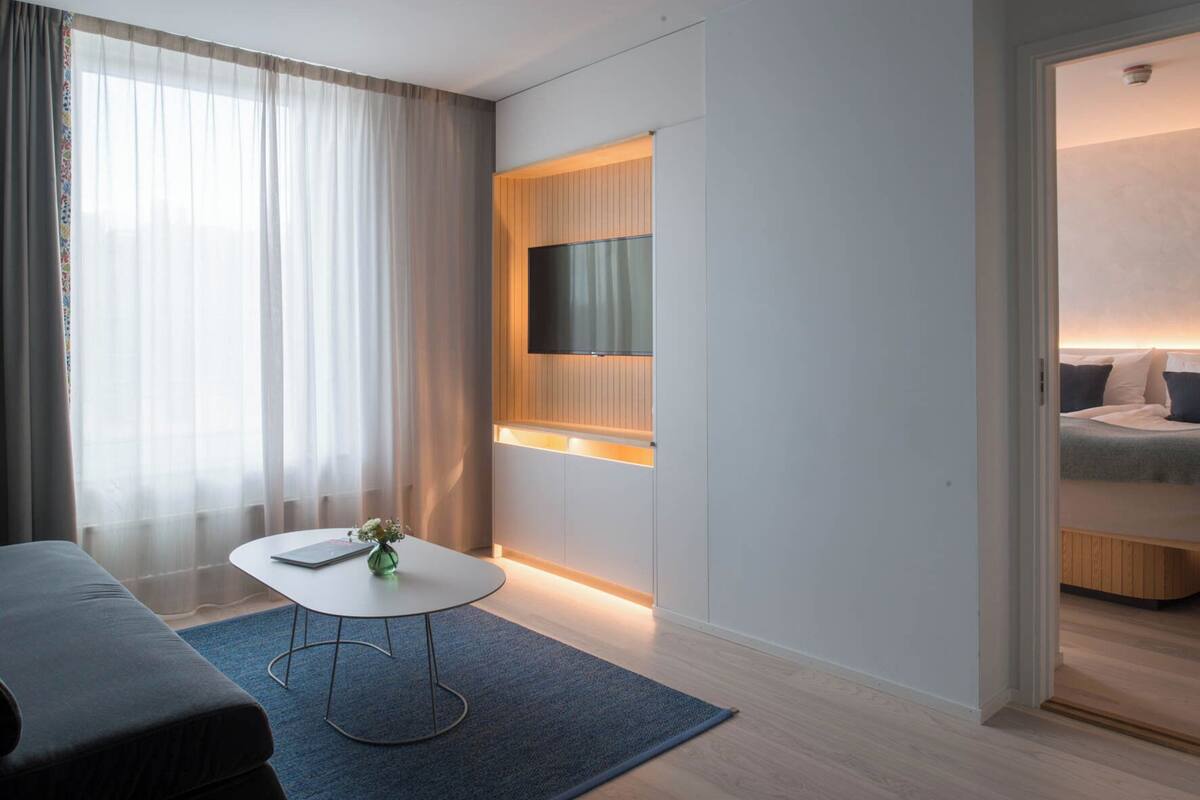 A modern hotel room with a bed, a small table with a vase, a TV mounted on a wooden panel, and a view of the sea through the window.