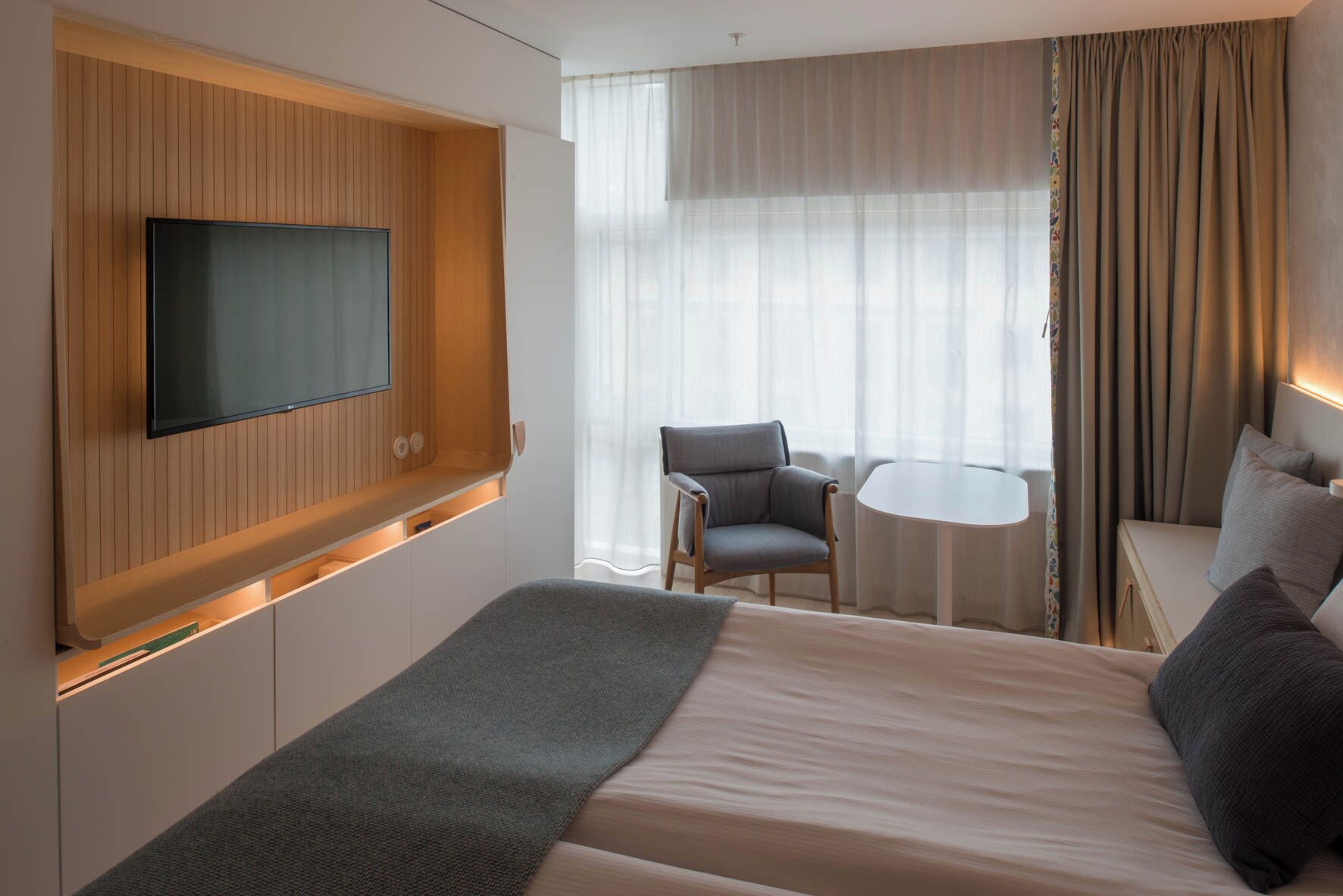 A modern hotel room with a large bed, a flat-screen TV mounted on a wooden panel wall, a small round table, a grey armchair, and a sofa with cushions.