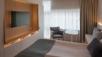Een moderne hotelkamer met een groot bed, een flatscreen televisie bevestigd aan een wand van houten panelen, een klein rond tafeltje, een grijze fauteuil en een bank met kussens.