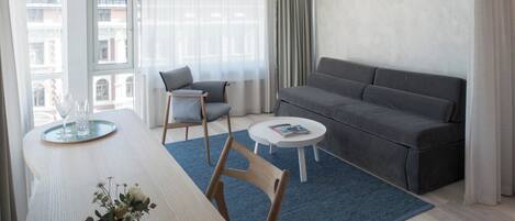 Una sala de estar moderna con un sofá gris, una mesita redonda, una mesa de comedor con sillas y vistas a edificios a través de ventanas amplias.