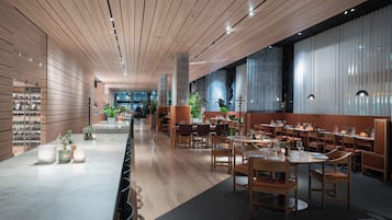 A modern restaurant interior with wooden ceiling panels, long dining tables, and chairs.