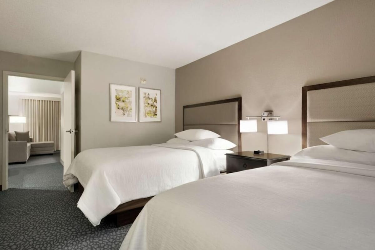 Una habitación de hotel con dos camas, una mesita de noche, una lámpara y una puerta que da a otra estancia.