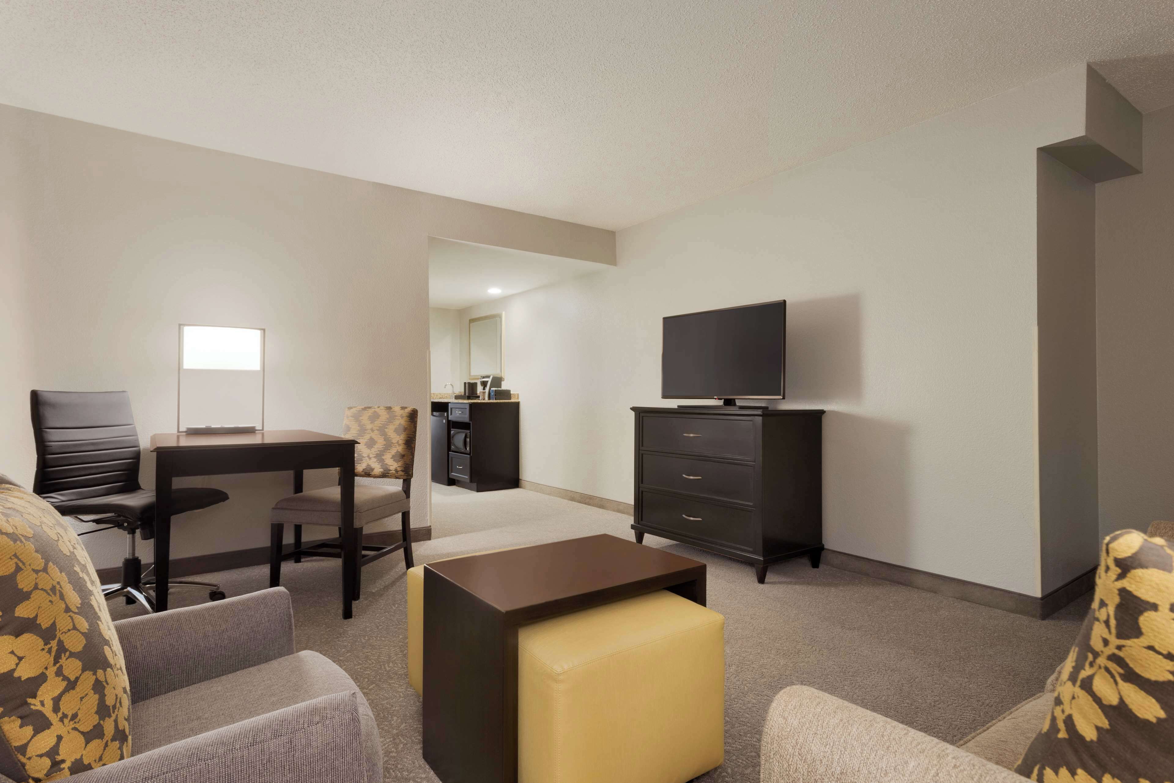 A modern hotel room with a flat-screen TV, a sofa, a coffee table, a dining table, and a small kitchenette.