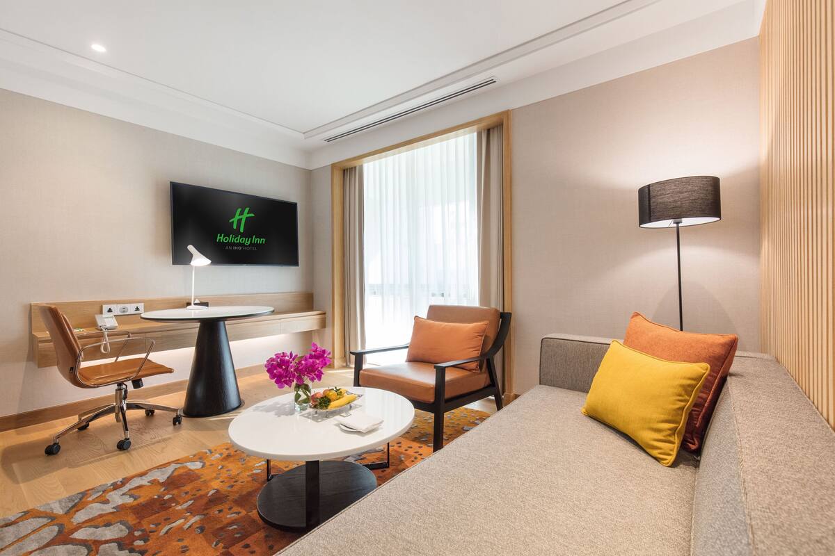 Una camera d'albergo moderna con televisione a schermo piatto, un divano, una sedia, un tavolino con un vaso di fiori e una lampada.