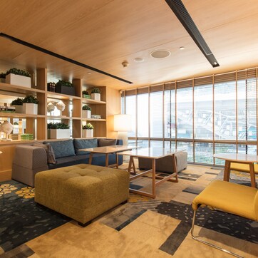 A modern living room with a sofa, ottoman, and chairs, a small table, shelves with plants, and a large window with a city view.