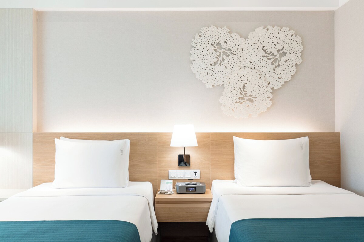 Hotellihuone, jossa on kaksi sänkyä, yöpöytä lampun kanssa, seinäkello ja koristeellinen seinäkoriste.