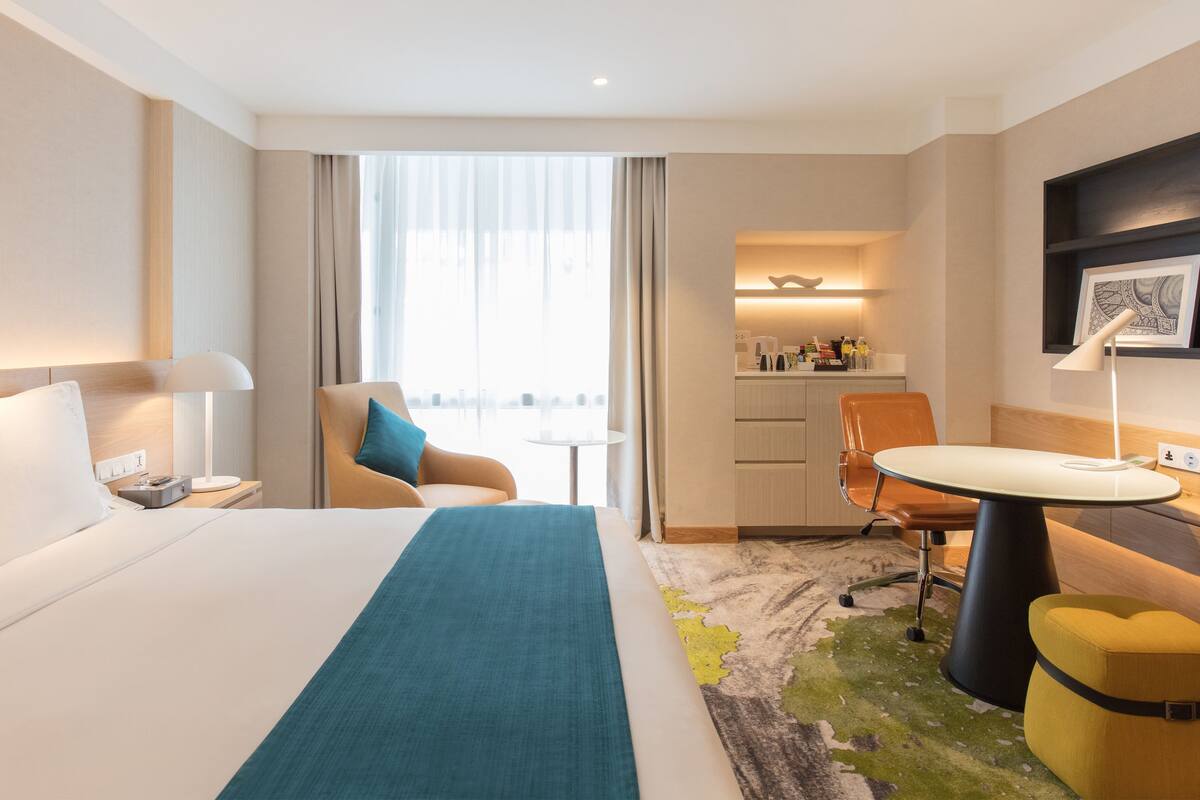 Una camera d'hotel con un letto, una scrivania, una sedia, un tavolino e vista sulla città tramite ampie finestre.