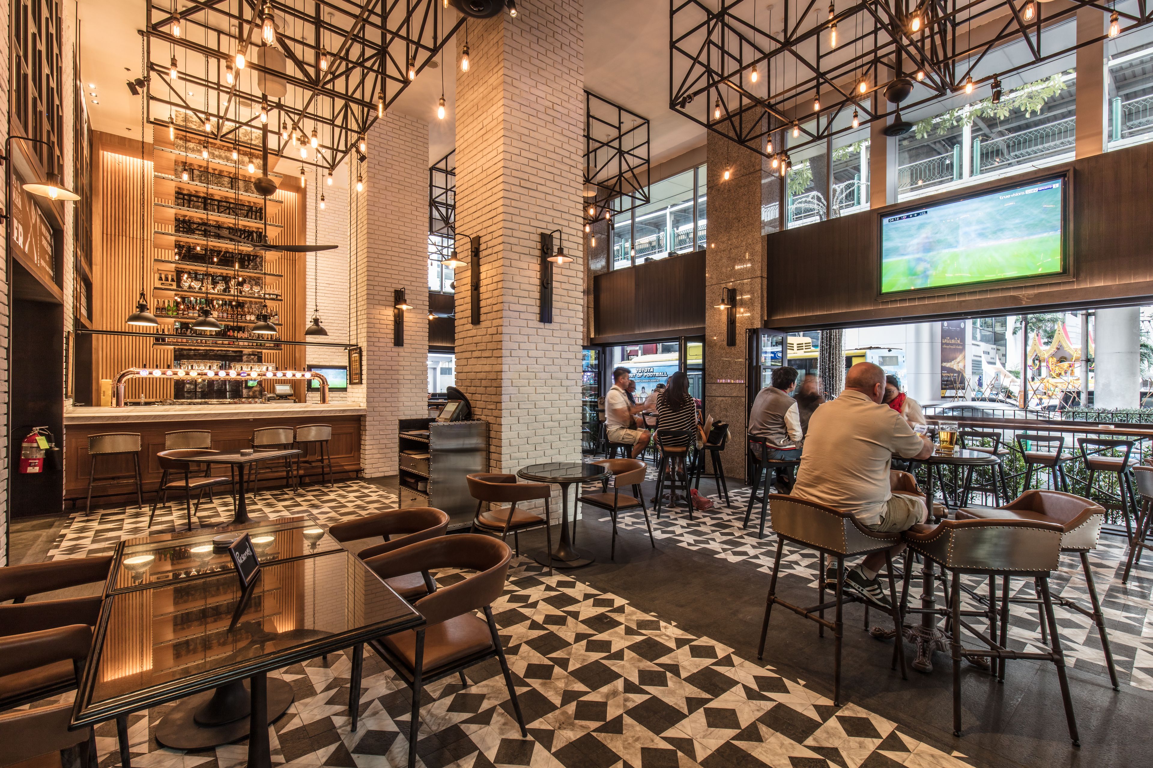 Un área de bar moderna con suelo a cuadros, taburetes y una pantalla grande mostrando un evento deportivo.