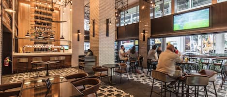 Un área de bar moderna con suelo a cuadros, taburetes y una pantalla grande mostrando un evento deportivo.